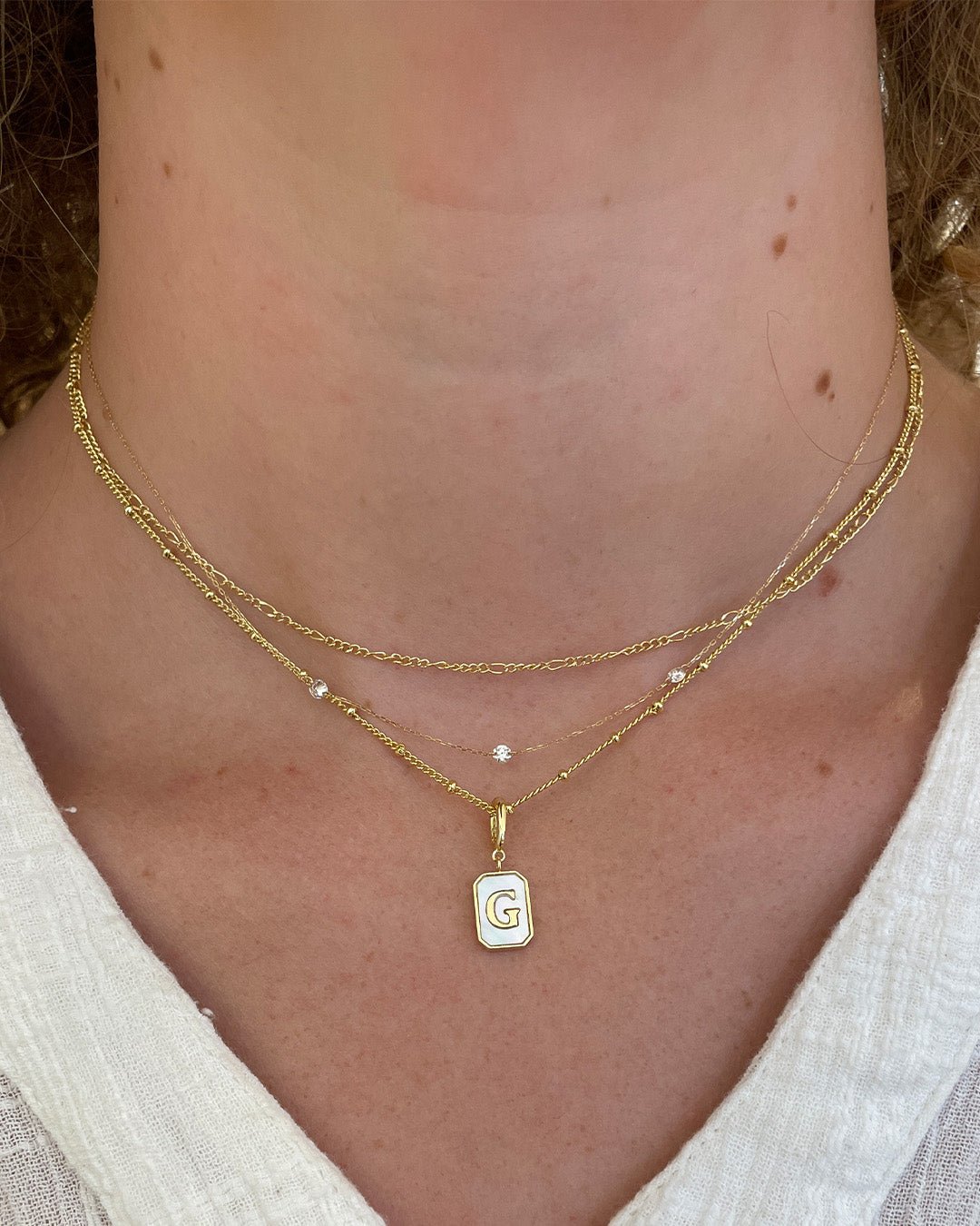 Woman wearing gold plated chain necklaces with an alphabet charm and a diamond trio necklace. 